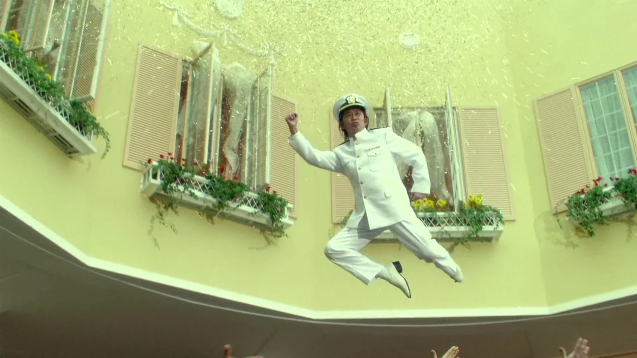 The Happiness of the Katakuris (2001) movie still: fullbody — A man in a white naval uniform leaps through the air in front of a yellow building…