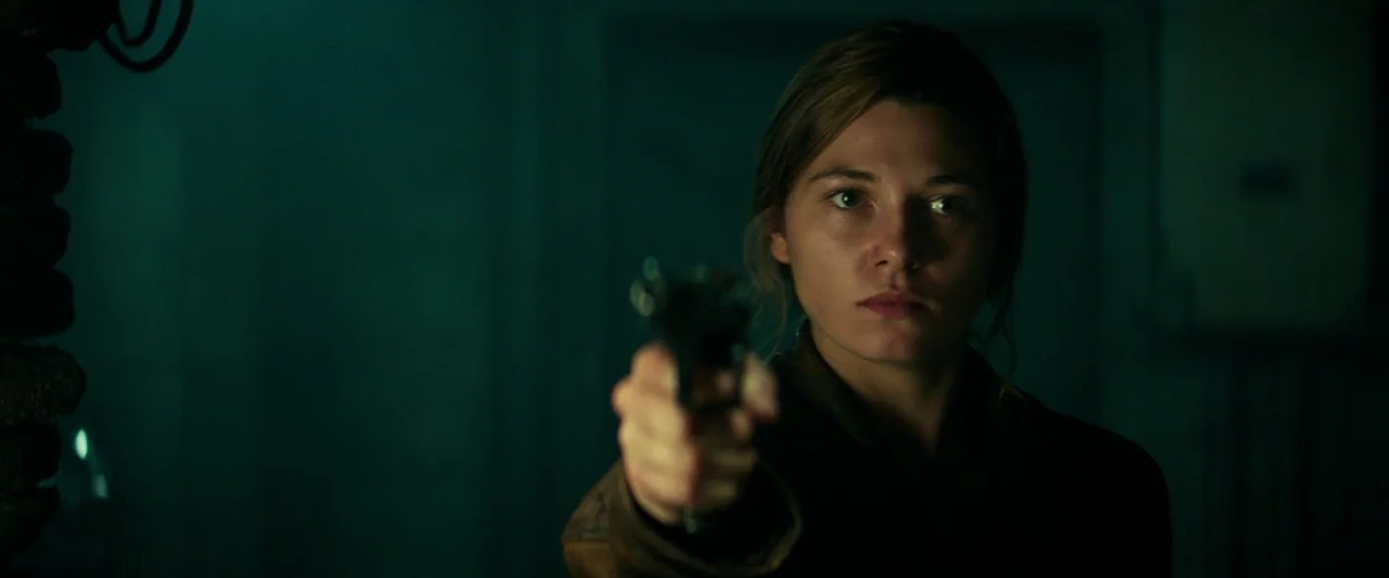 Overlord (2018) movie still: medium — A dimly lit room with dark walls and indistinct architectural details. A woman stands in the center, aiming a…