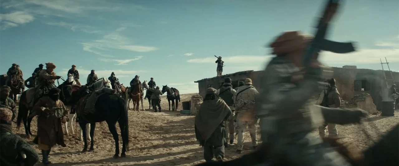 12 Strong (2018) movie still: wide — A group of armed men gather in a dusty desert village with mud structures. Horses are tethered nearby while men…