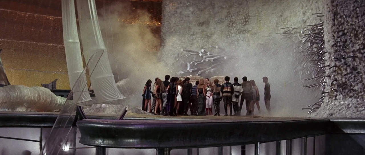 Barbarella (1968) movie still: wide — A group stands on a platform surrounded by metallic textures and billowing white fabric sheets. The figures…