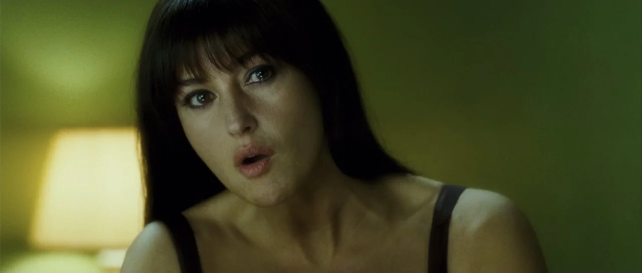 Shoot 'Em Up (2007) movie still: closeup — An interior bedroom with green walls lit by a dim bedside lamp. A woman with dark hair looks towards the…