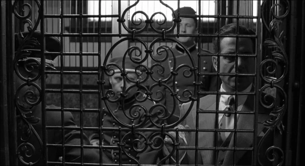 The Man Who Wasn’t There (2001) movie still: medium — A metal cage elevator interior with an ornate decorative wrought iron grate barrier. Three men…