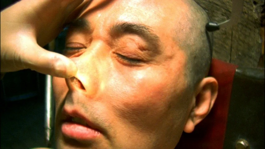 Save The Green Planet! (2003) movie still: closeup — A close-up view shows a man with a shaved head in a sterile environment. A hand grips the…
