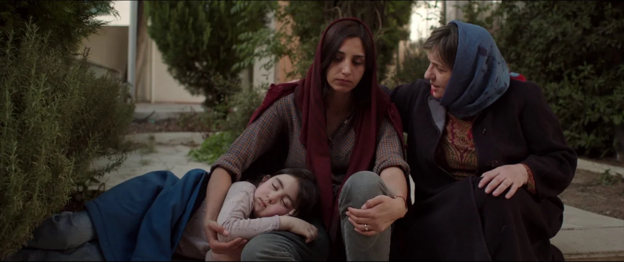 Under The Shadow (2016) movie still: medium — A quiet outdoor courtyard features concrete paving and dense green shrubs in the background. Two adult…