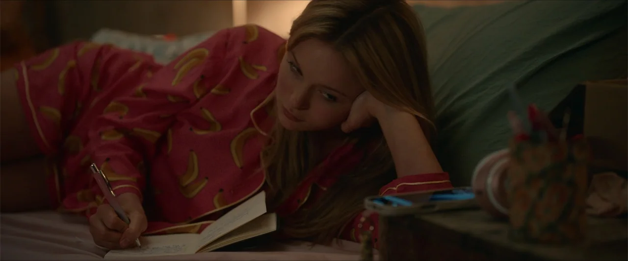 Love Me Love Me (2026) movie still: medium — A dimly lit room with a person resting on a bed covered in green linens. A young woman in patterned…