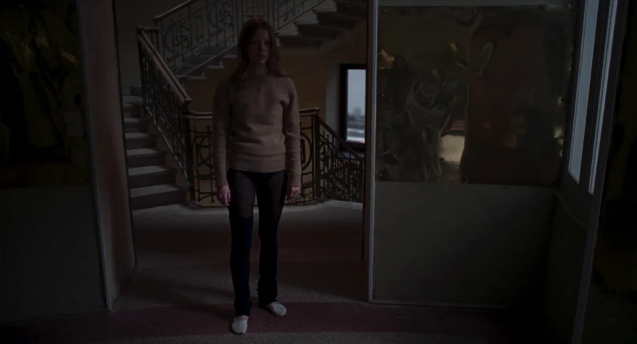 Suspiria (2018) movie still: fullbody — A dark stairwell landing features a wooden banister and reflective decorative wall panels. A young woman…