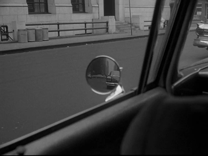 Blast Of Silence (1961) movie still: medium — A view from inside a car looking out at a city street and sidewalk. A side mirror reflects a trailing…