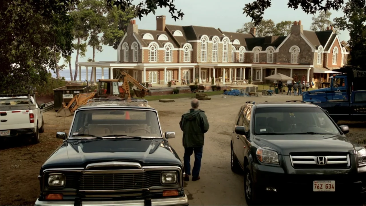 Clear History (2013) movie still: fullbody — A construction site sits before a large multi-story brick estate near the water. An older man stands…