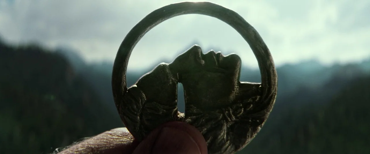 The Lone Ranger (2013) movie still: closeup — A dark metallic circular pendant with carved profiles of two figures is held up. The background…