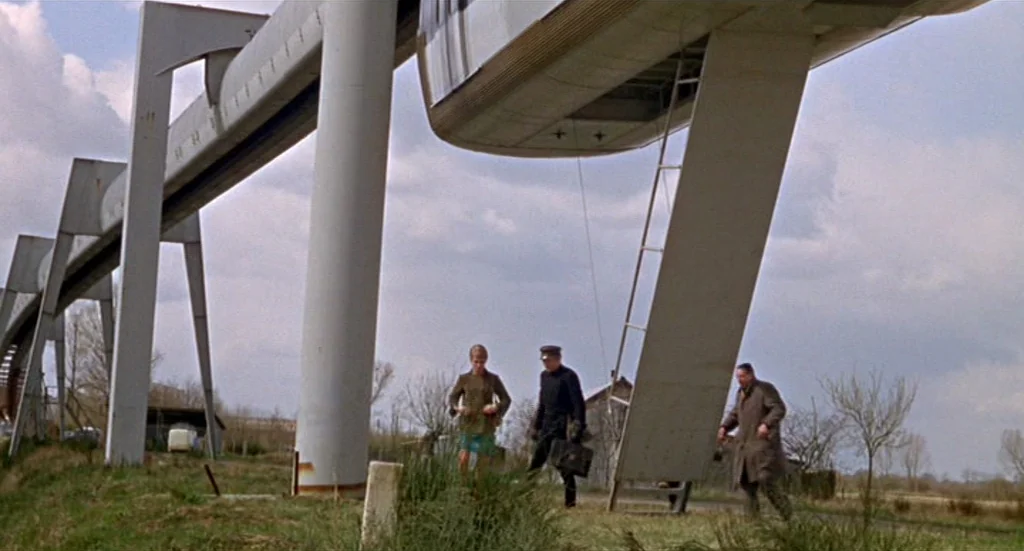 Fahrenheit 451 (1966) movie still: wide — An industrial monorail track sits on elevated concrete pillars over a grassy field. A woman, a man in…