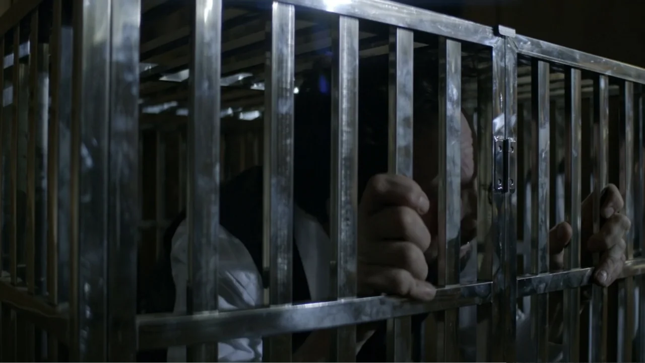 The Collective (2023) movie still: medium — A man stands trapped behind the cold vertical metal bars of a locked cage. He grips the metallic frame…