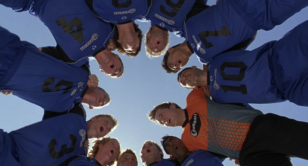 She's the Man (2006) movie still: wide — A group of soccer players huddle in a circle on a bright sunny day. They are wearing athletic jerseys and…