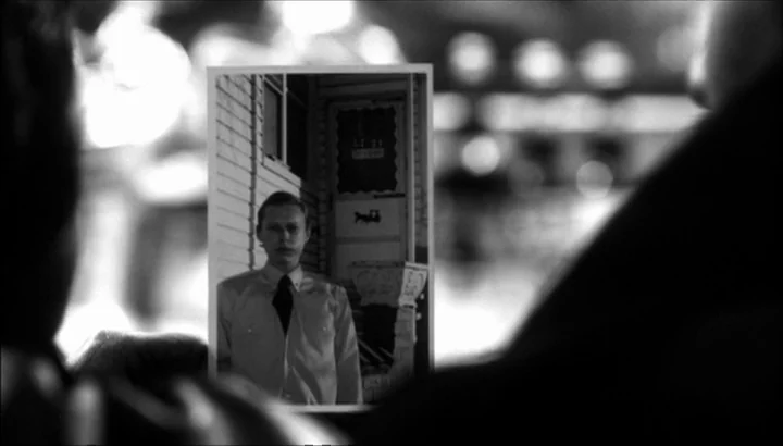 The American Astronaut (2001) movie still: closeup — A person holds a black-and-white photograph of a man in front of a door. The foreground is…