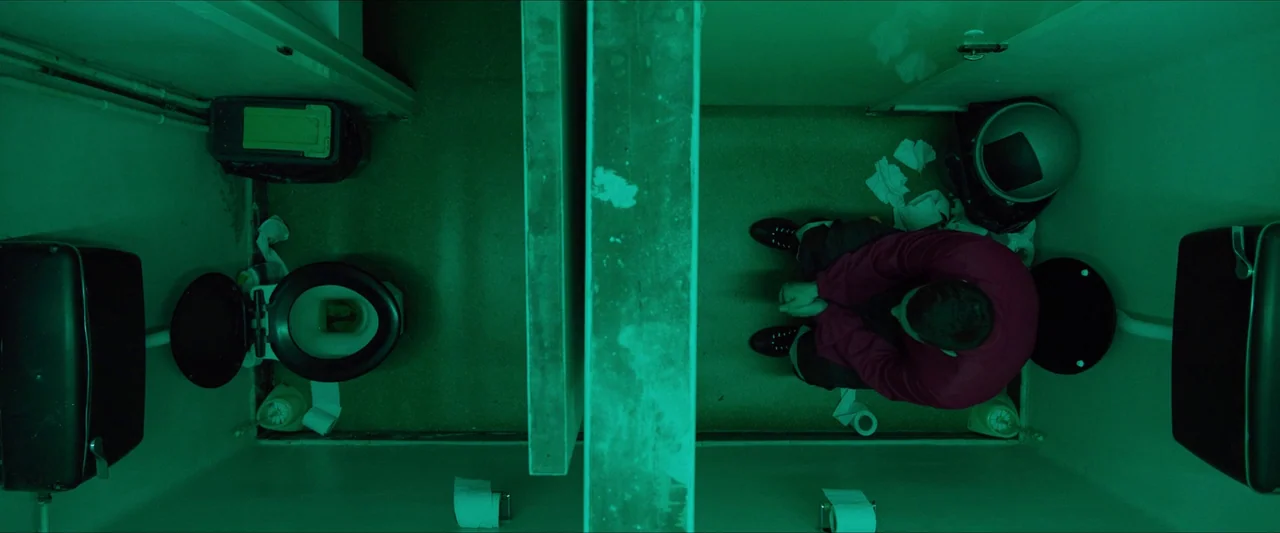 Kneecap (2024) movie still: wide — A top-down view shows two cramped stalls divided by a solid concrete-like partition. A lone individual sits on…