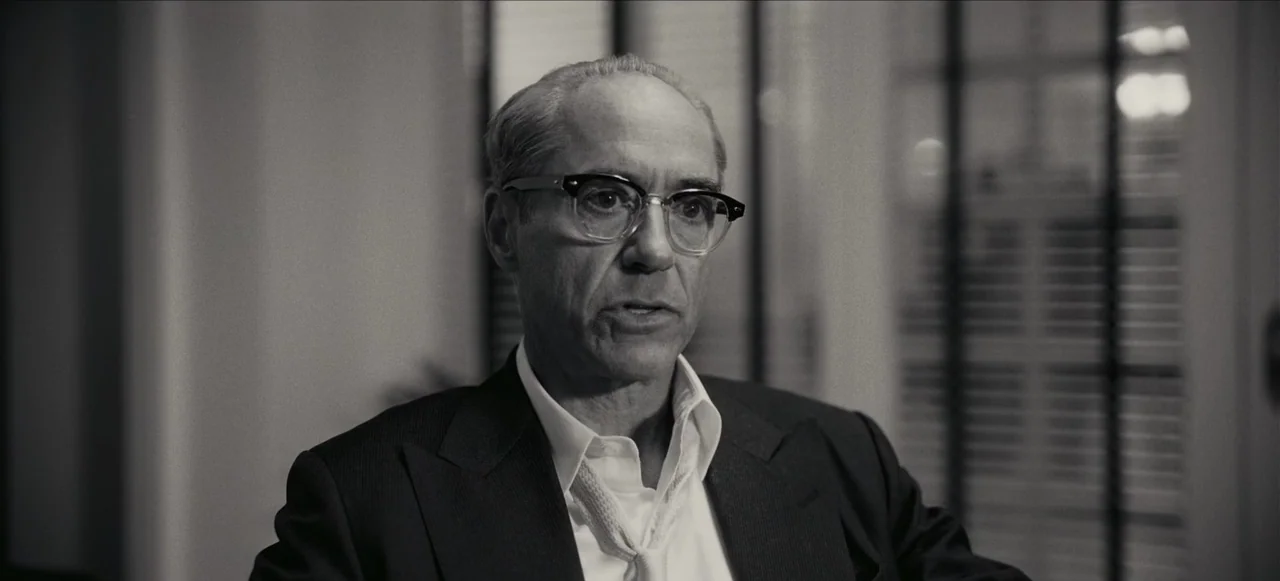Oppenheimer (2023) movie still: closeup — An interior office with venetian blinds visible in the blurred background. A middle-aged man in a suit is…