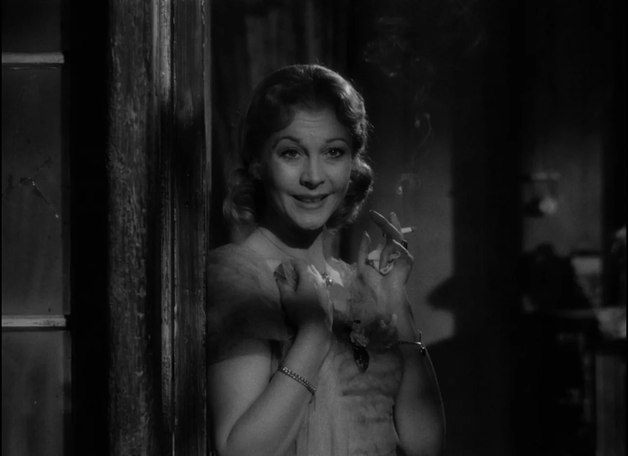 A Streetcar Named Desire (1951) movie still: medium — A dimly lit interior space with a wooden door frame and dark shadows. A woman leans against…