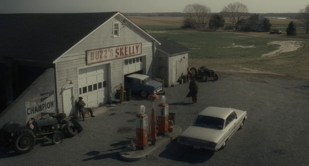 Inside Llewyn Davis (2013) movie still: wide — A rural, weathered gas station sits beside a field under a clear sky. Two men are present in the…