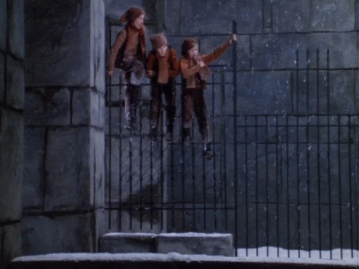 The Magic Flute (1975) movie still: wide — Three children in matching orange-brown clothing climb over an iron fence at night. A large grey stone…