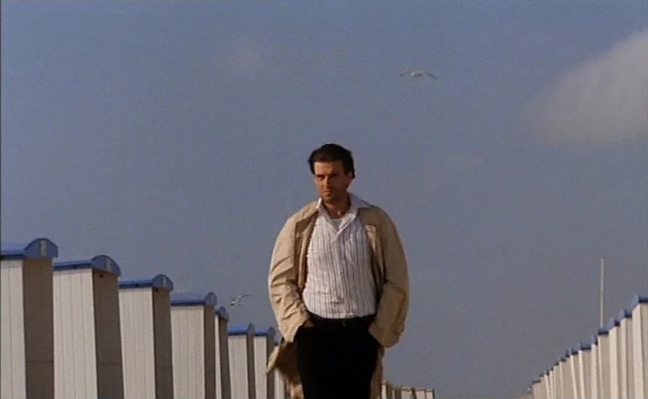 The Fourth Man (1983) movie still: fullbody — A beach boardwalk lined with rows of identical white changing stalls under a cloudy sky. A man walks…