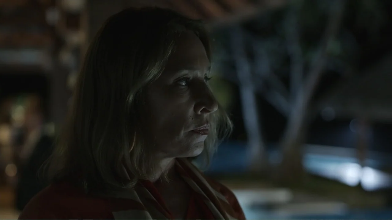 Unknown #1 movie still: closeup — An outdoor patio at night features blurred dark foliage and light patches in the background. A woman in a red top…