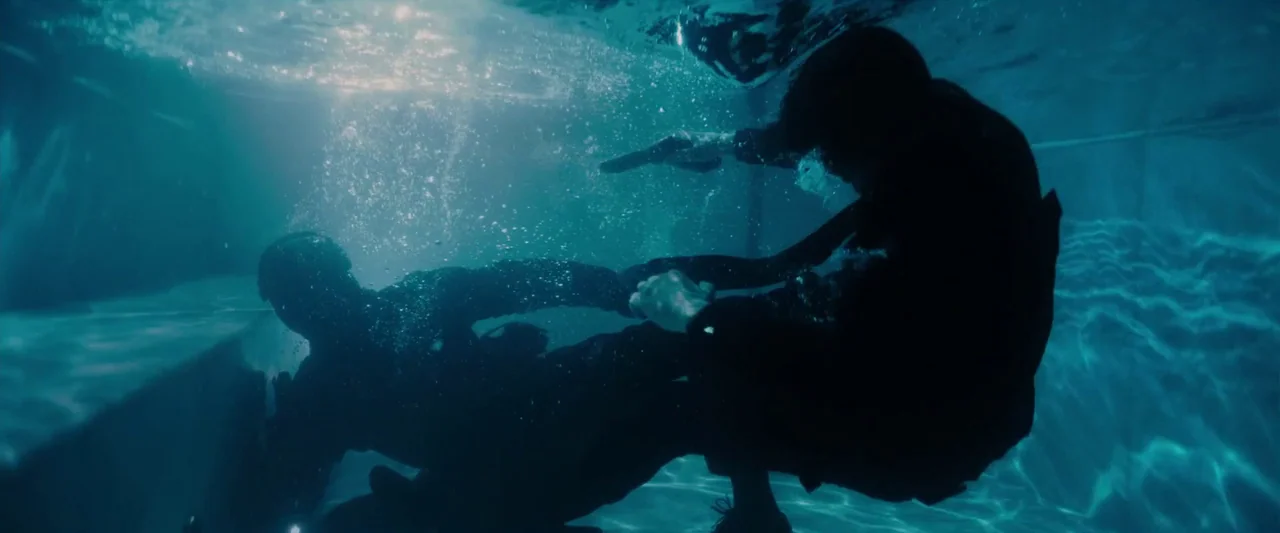 John Wick: Chapter 3 – Parabellum (2019) movie still: wide — Two figures are submerged underwater in a pool during the daytime. One person holds a…