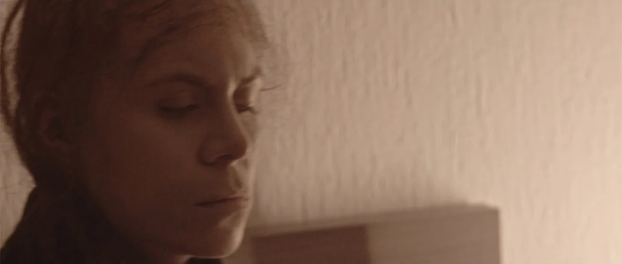 Amor Mutante (2019) movie still: closeup — A plain interior wall with a subtle stucco texture provides a backdrop. A young woman leans her head…