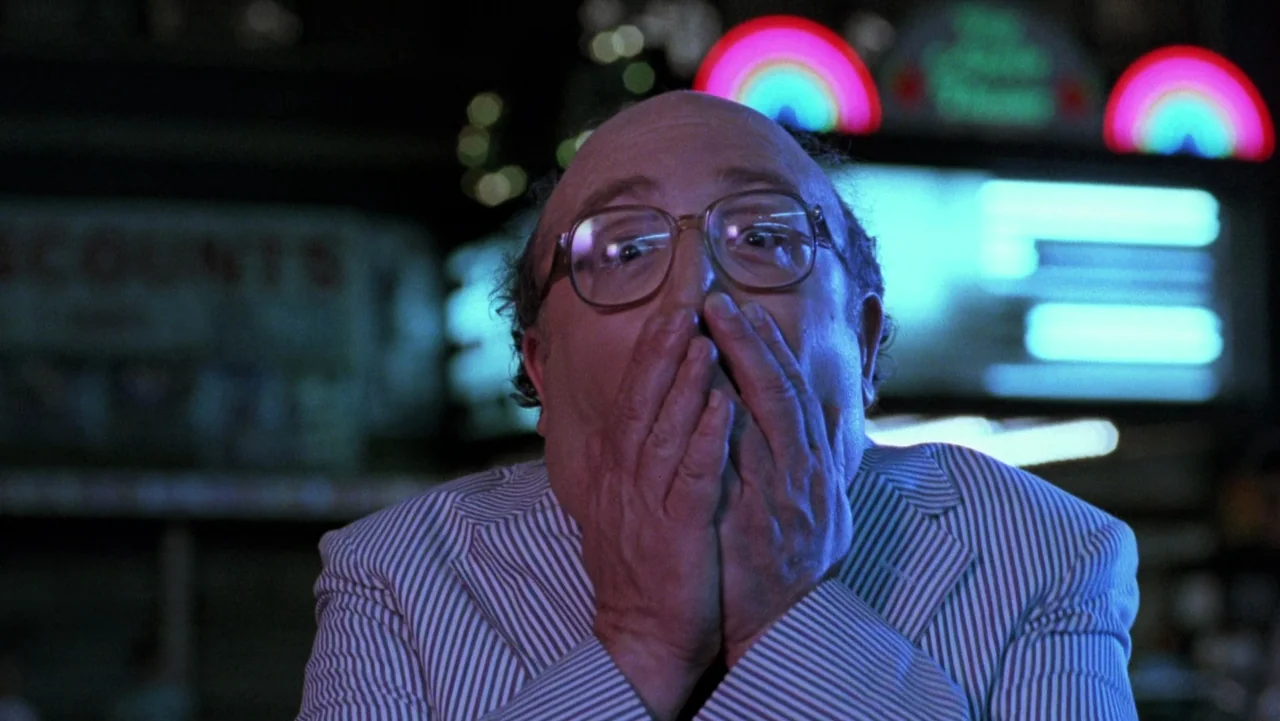 Frankenhooker (1990) movie still: closeup — An urban setting at night features neon signage with blurred rainbow patterns in back. A bald man…