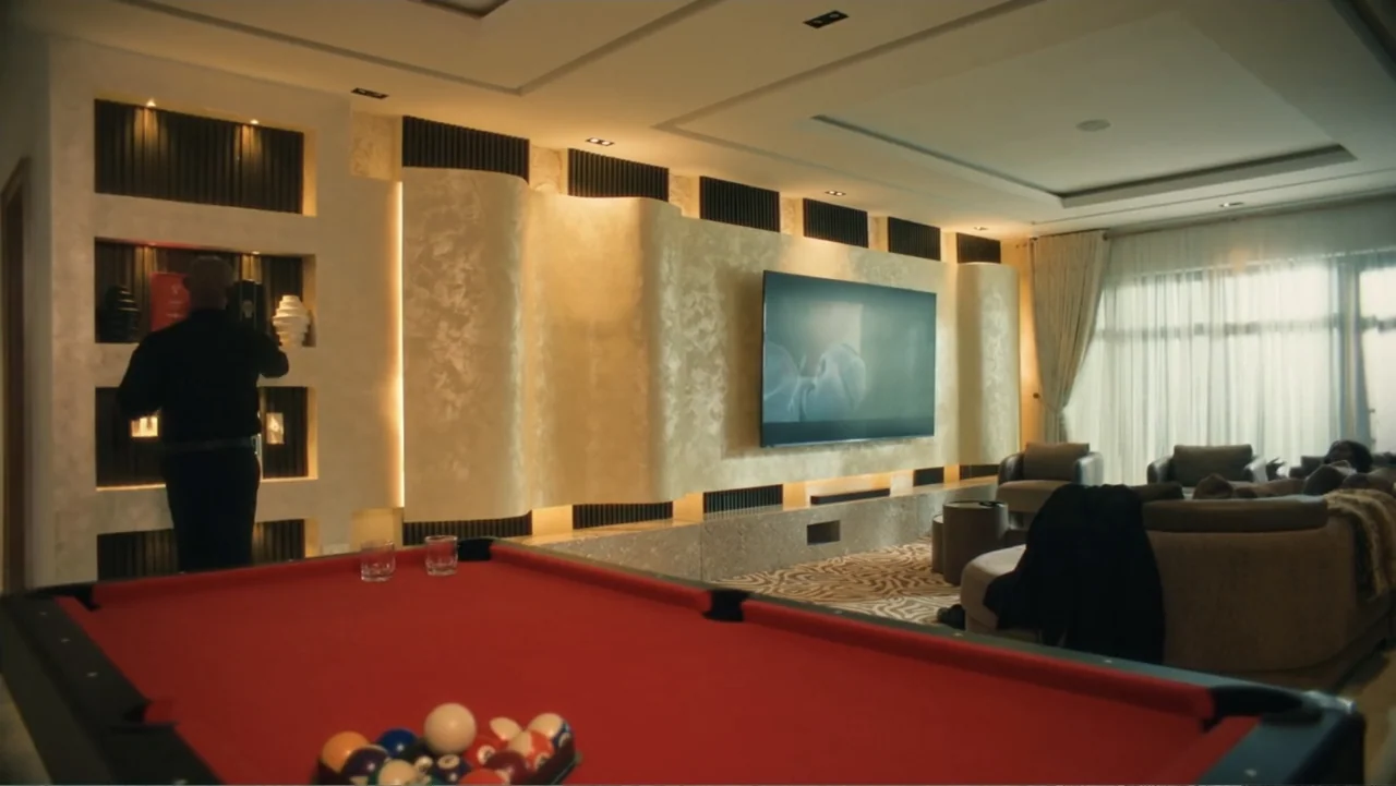 SIN: Blackout (2025) movie still: wide — A modern luxury living room features a large television wall with curved gold panels. A man in black stands…