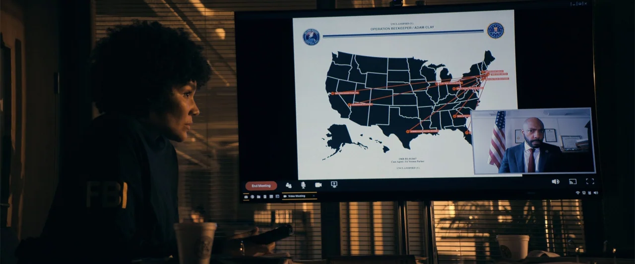 The Beekeeper (2024) movie still: medium — A dimly lit office features a large computer screen displaying an FBI map and a video call. A woman with…