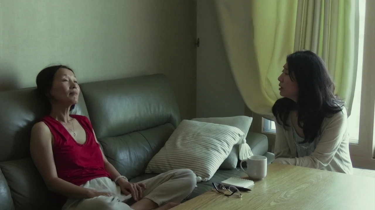 In Front Of Your Face (2021) movie still: medium — An interior living room features a green leather couch near a large window. Two women sit on the…