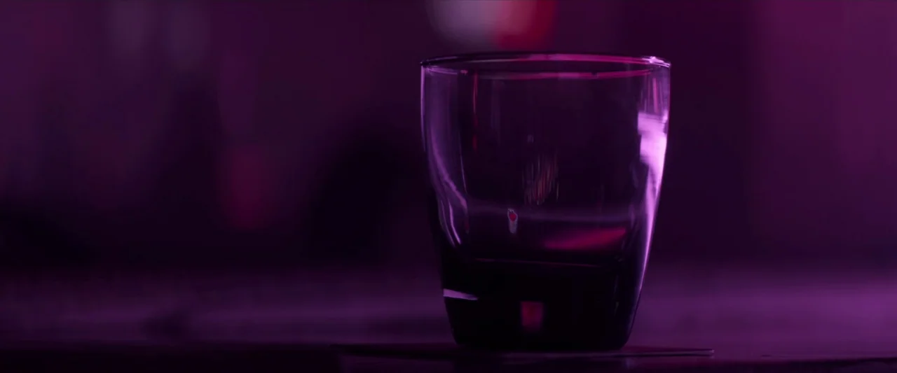 The Silencing (2020) movie still: closeup — A glass sits on a dark surface in a dimly lit, purple-hued environment. The background is completely out…