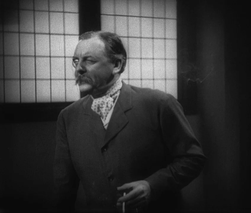 The Threepenny Opera (1931) movie still: medium — A man in a dark suit stands before a window with a grid pattern. He is positioned indoors, holding…