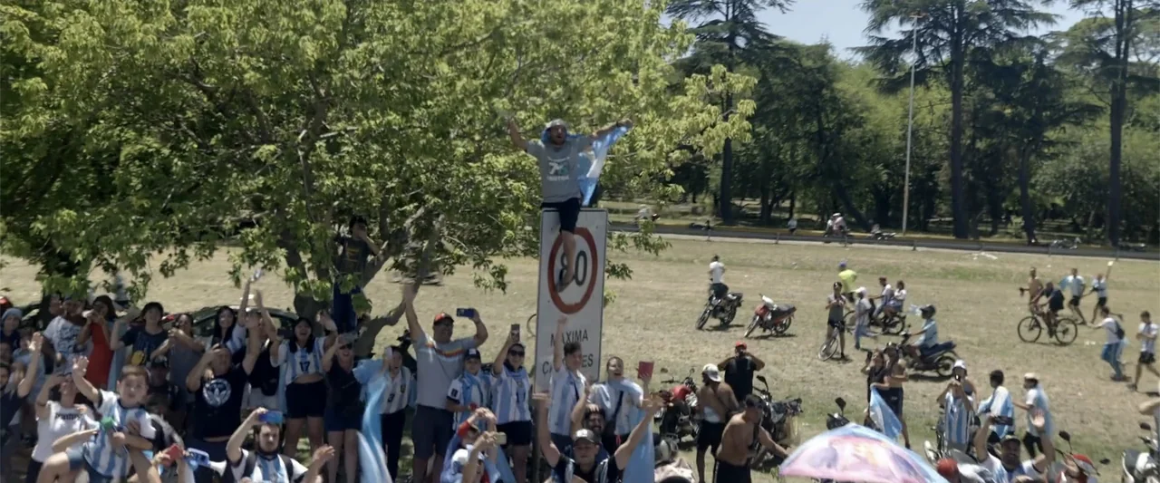 I Choose to Believe (2023) movie still: wide — A sunny public park features a large crowd of people wearing football jerseys. A man stands balanced…