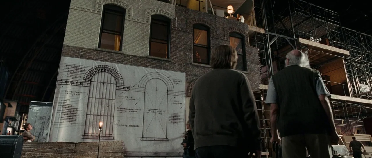 Synecdoche, New York (2008) movie still: wide — A vast indoor film studio features a multi-story building facade under active construction. Two men…