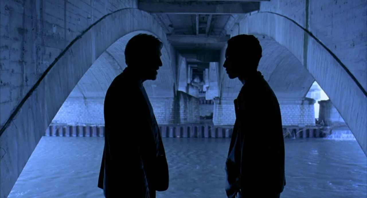 The Good Thief (2002) movie still: fullbody — A dark concrete underpass spans a river with large arches on both sides. Two men stand facing each…