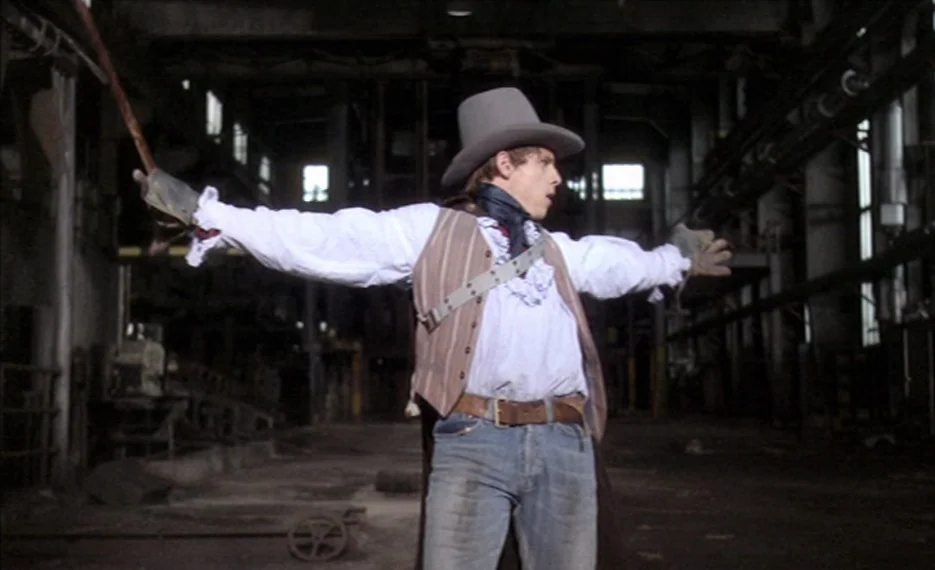 Dear Wendy (2005) movie still: medium — A dark, cavernous industrial space features exposed metal structures and concrete floor surfaces. A young…