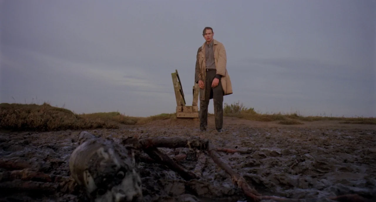 Possum (2018) movie still: wide — An overcast, murky mudflat stretches under a dim, grey twilight sky. A man stands silently in the muck, staring…