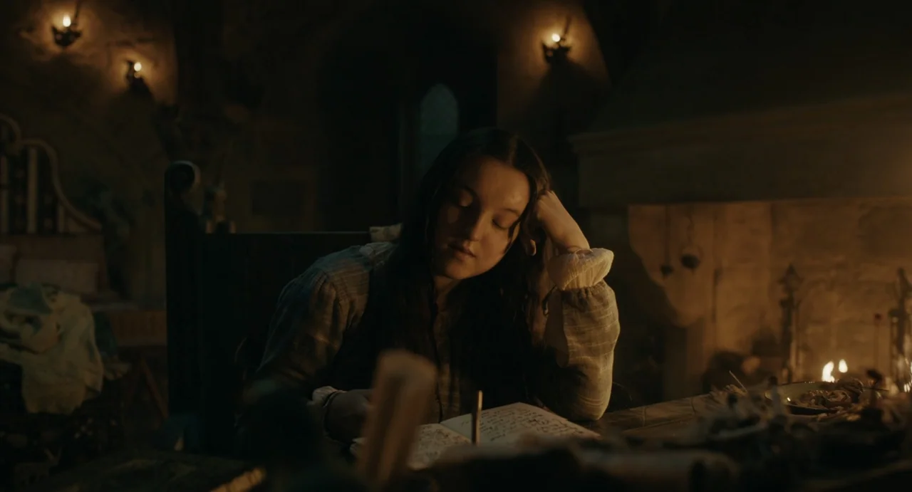 Catherine Called Birdy (2022) movie still: medium — A dimly lit room with heavy stone walls and a fireplace burning in the background. A young woman…