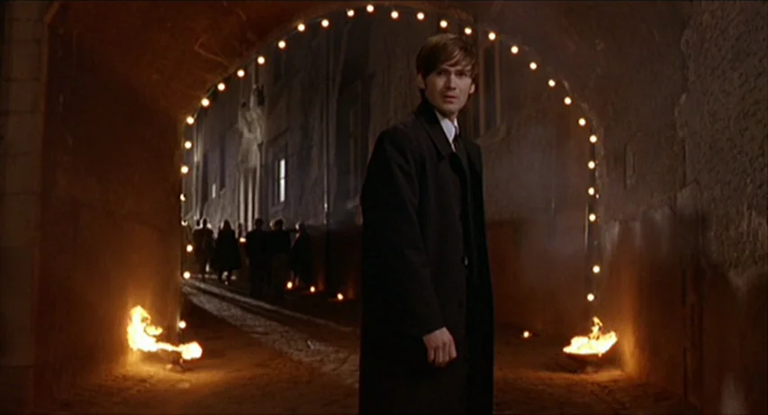 CQ (2001) movie still: medium — A stone tunnel archway features a string of small lights and fire bowls. A man stands in the foreground wearing a…