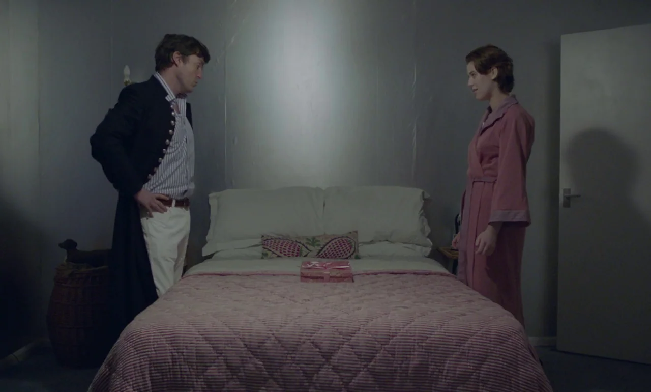 The Souvenir (2019) movie still: fullbody — A spartan bedroom features plain grey walls and a bed with a pink quilt. A man in a dark coat and a…