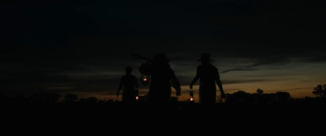 Mudbound (2017) movie still: wide — Three figures walk across an open, dark landscape during the final moments of sunset. They carry small handheld…