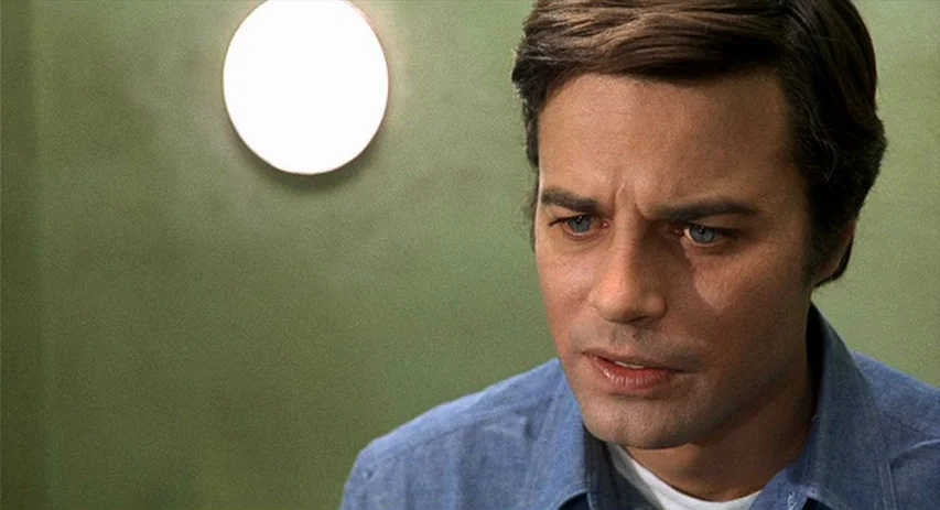 Perversion Story (1969) movie still: closeup — A man stands in front of a plain olive-green wall with a glowing circular light. He gazes downward…