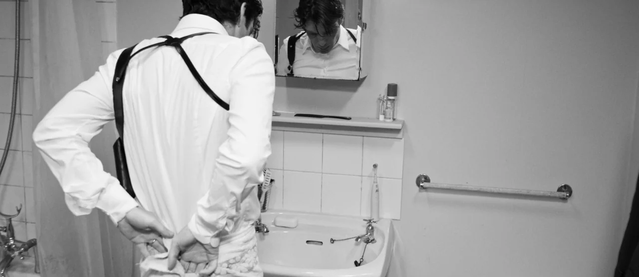 The Party (2017) movie still: medium — A utilitarian bathroom contains a white sink, tile backsplash, and a medicine cabinet. A man in a dress shirt…