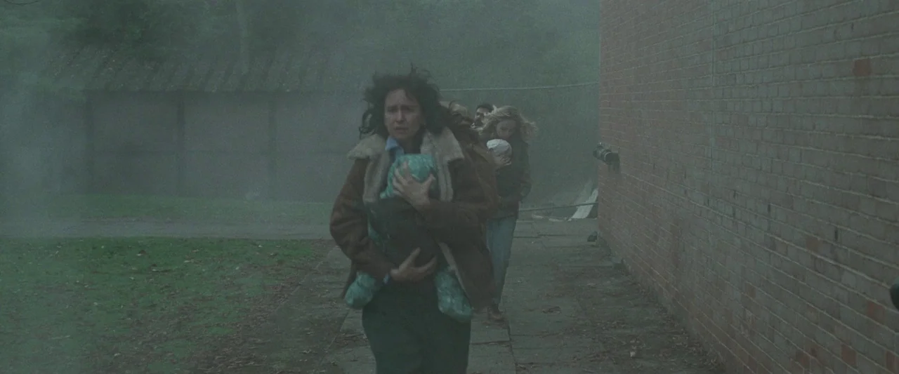 The End We Start From (2023) movie still: fullbody — A misty outdoor area with a brick wall and paved path beside grass. A woman with disheveled…