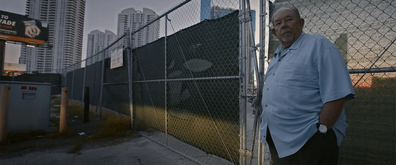 The Kleptocrats (2018) movie still: medium — An outdoor construction site is bordered by a tall chain-link fence with privacy netting. An elderly…