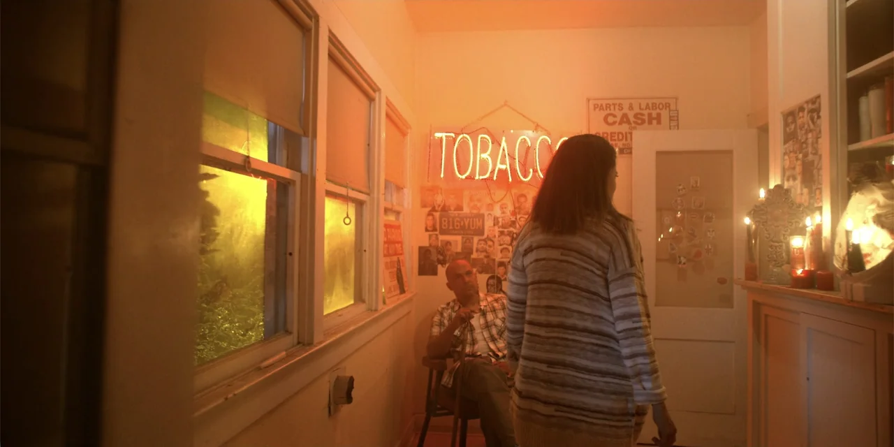 7th & Union (2021) movie still: medium — A narrow sunroom with cream-colored walls and a wall decorated with photographs and posters. A woman in a…