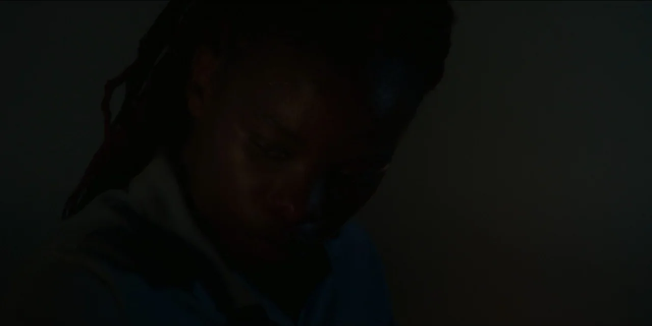 The Hunted (2024) movie still: closeup — A dimly lit indoor space with low light levels obscures the background details. A person with locs and a…