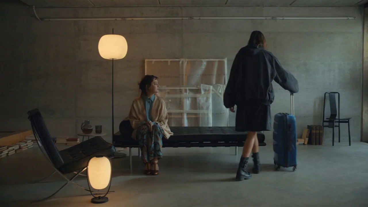 Amanda (2022) movie still: wide — A minimalist room with raw concrete walls, a black leather bench, and floor lamps. One woman sits on the bench…