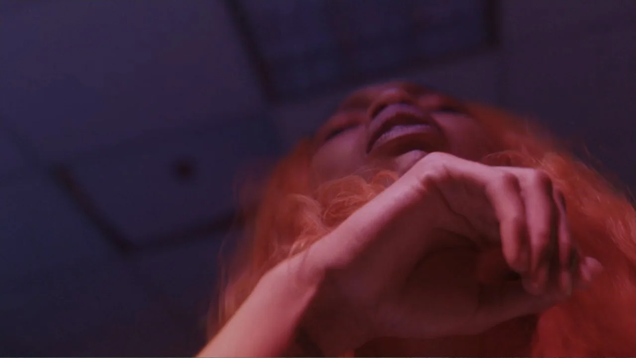 The African Desperate (2022) movie still: closeup — An interior space bathed in deep purple and magenta light. A woman with bright orange hair looks…