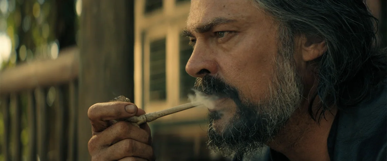 The Bluff (2026) movie still: closeup — A wooden porch area features blurred background vertical slats in soft golden light. A bearded man in his…